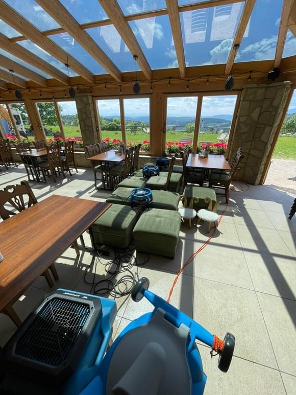 Proces szybkiego suszenia wypranej tapicerki w restauracji z widokiem na góry. Na zielonych pufach ustawione są profesjonalne osuszacze, które skracają czas schnięcia mebli w obiektach komercyjnych.